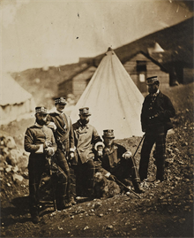 Black and white photograph of bearded soldiers in front of a tent and huts.