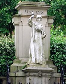 Nightingale statue Derby. Set back from a road with trees/vegetation in the background. Nightingale shown standing, holding a lamp at head height, in front of the words 'Fiat Lux'.