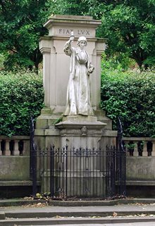 Nightingale statue Derby. Set back from a road with trees/vegetation in the background. Nightingale shown standing, holding a lamp at head height, in front of the words 'Fiat Lux'.